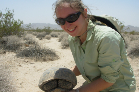 Dr Chava Weitzman wearing sunglasses holding a tortoise, in arid-looking country with small shrubs in the background