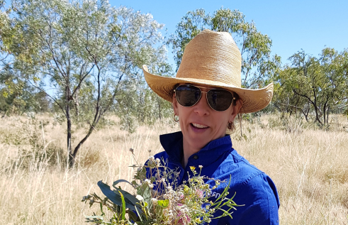 Dr Donna Lewis head and shoulders, wearing hat and sunglasses and holding a bunch of native flowers and leaves, with grass and trees in background