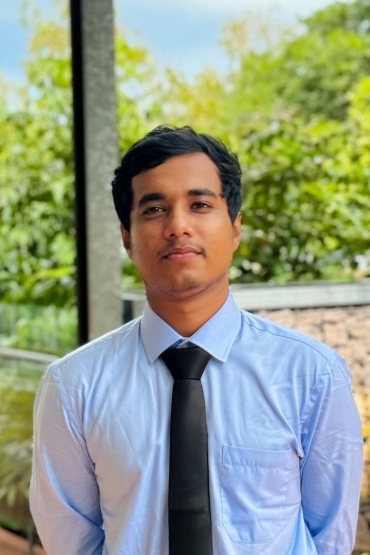 A portrait of a brown man standing in front of a brick wall, wearing a blue shirt, and a black tie.