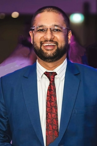 A portrait of a brown-bearded man wearing glasses, a blue suit, and a red tie, smiling.