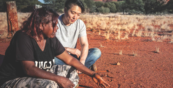 Indigenous woman explaining a figure drawn in the dirt to a man who is sitting with her and listening