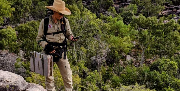 Person wearing hat standing on rocks looking at GPS, with trees in background