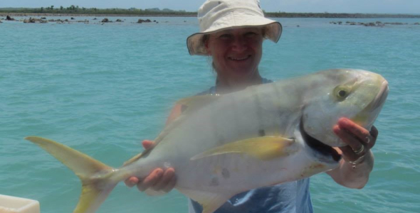 person wearing hat, standing in boat, holding large fish, with water in the background