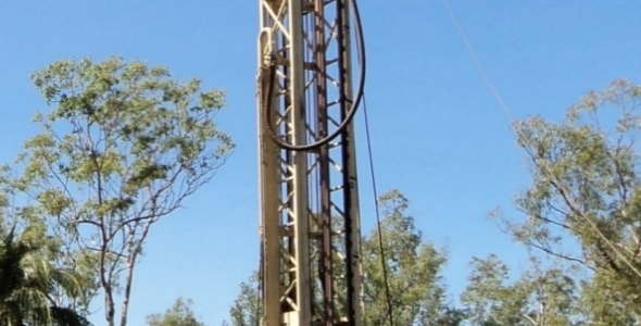 Tall drill rig on back of truck, with person in hard had and hi-vis working at the bottom, with trees in the background