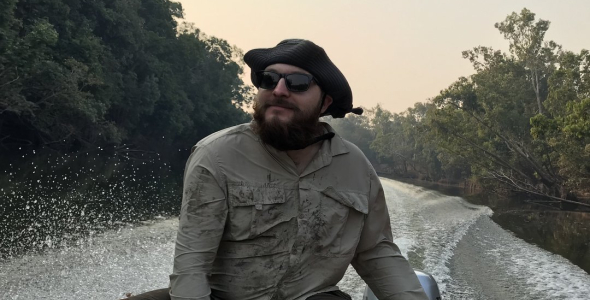 Cameron Baker wearing sunglasses and hat, steering a boat on a river lined with forest, with wake behind