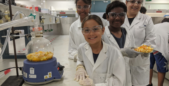 three children and an adult wearing white coats and protective glasses, in a laboratory