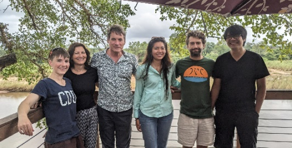 six people standing in a row, posing for photo, with vegetation in background