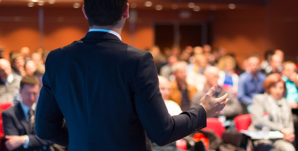 In an auditorium, a presenter in a suit addresses an audience