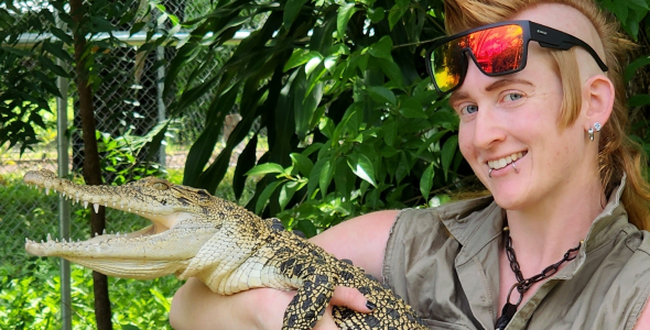 Person with mohawk hairstyle, with sunglasses on their forehead, holding crocodile about the size of a medium sized dog, with green leafy background