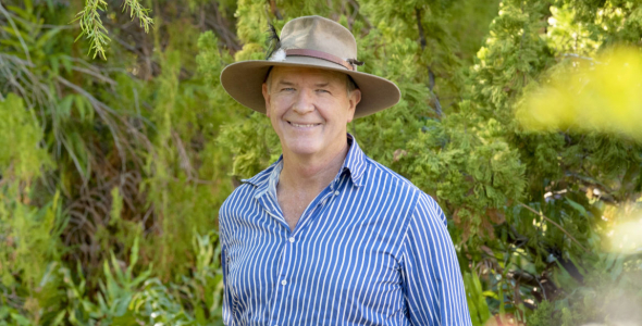 Professor Stephen Garnett head and shoulders, wearing blue and white striped shirt and an Akubra hat, with green leafy vegetation in background