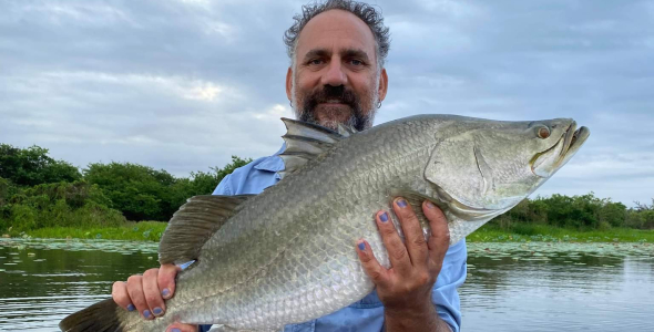 Colton Perna holding a large fish, standing in a boat on water, with bushes and other plants on the bank behind