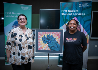 Caucasian woman wearing glasses with a proud smile on her face standing next to an artwork of a blue cockatoo with a young Aboriginal woman on the right also with a big proud smile on her face. 