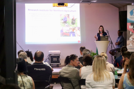 people sitting with backs to camera, Maxine Piggott standing on stage speaking, with presentation slide projected onto screen next to her