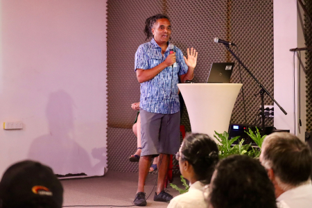 Sunil Kadri standing by a round lectern with a laptop on it, with one hand raised and a microphone in the other, speaking to people with their backs to the camera speaking to