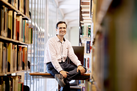 Man sitting on desk surrounded by books in a light filled room.