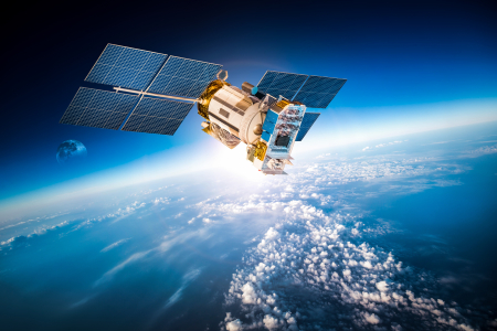 satellite with four solar panels on both sides, above cloudy blue atmosphere, against sky ranging in colour from black through light blue to white near the horizon, with partial moon visible in background