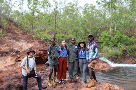 Six people looking at camera smiling standing on a rock in front of a waterfall. 