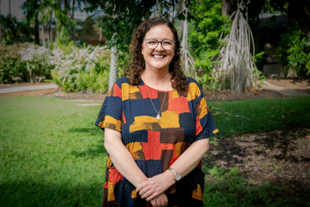 Woman wearing glasses smiling at camera in a colourful top. She is standing in a garden. 