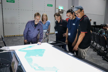 5 people standing around a large table with a map projected onto it