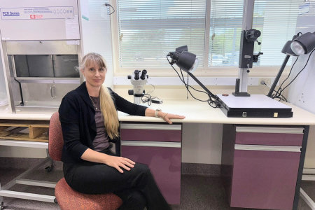 Woman seated in front of laboratory equipment looking directly at camera. She has long blonde hair that is styled in a ponytail.  