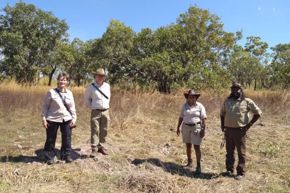 four people standing in an open space with trees and grass in the background