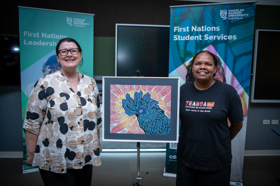 Caucasian woman wearing glasses with a proud smile on her face standing next to an artwork of a blue cockatoo with a young Aboriginal woman on the right also with a big proud smile on her face. 