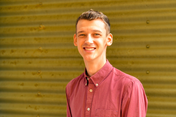 Sam Williams - young man smiling and wearing a light red button up shirt smiling in front of corrugated iron wall