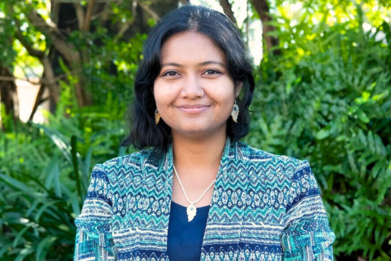 Staff portrait - smiling woman with short black hair and stripey blue jacket standing against foliage