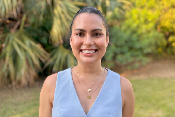 Sharna Motlap - a young indigenous woman smiling with light blue vest in front of palm trees