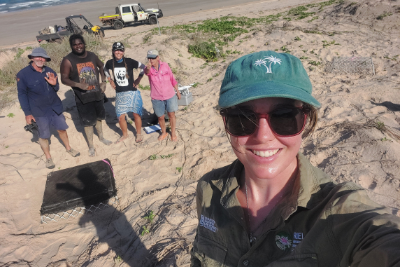 A young woman takes a selfie with two women and two men behind her. They are on a beach. 