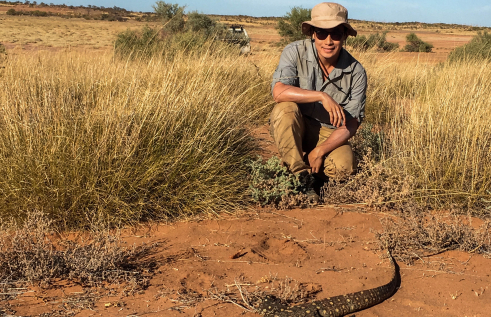 Dr Nicholas Wu, wearing a hat and sunglasses, crouching near some clumps of grass, with a large lizaard on bare soil in the foreground