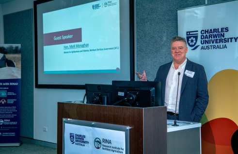 Man in a dark blue jacket gestures behind a lectern, with Charles Darwin University banner behind him and  "Guest speaker, Hon Mark Monaghan, Minister for Agribusiness and Fisheries, Northern Territory Government (NTG)" on screen to his right.