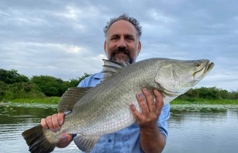 Colton Perna holding a large fish, standing in a boat on water, with bushes and other plants on the bank behind