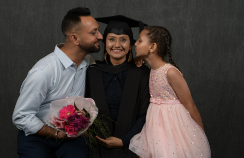 Indian man wearing light blue collared shirt, Indian woman wearing black academic gown and hat and their young daughter in pink dress with tutu.