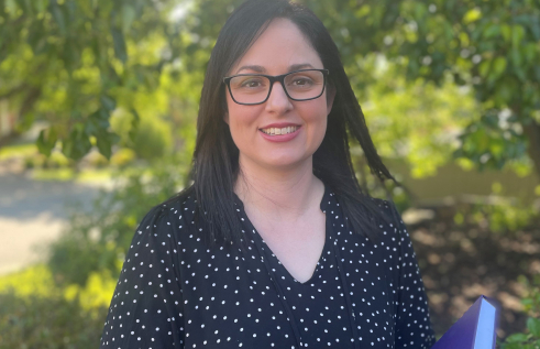 Caucasian woman holding a book standing in front of a tree with lots of green leaves. She has brown hair and is wearing optical glasses and smiling.