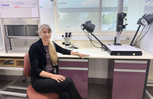 Woman seated in front of laboratory equipment looking directly at camera. She has long blonde hair that is styled in a ponytail.  