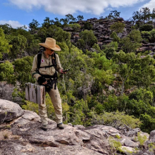 Person wearing hat standing on rocks looking at GPS, with trees in background