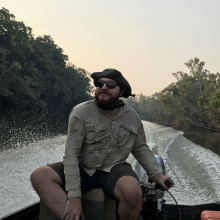 Cameron Baker steering a boat on a river lined with forest