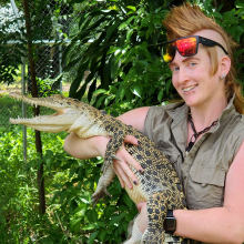 Person with mohawk hairstyle, with sunglasses on their forehead, holding crocodile about the size of a medium sized dog, with green leafy background