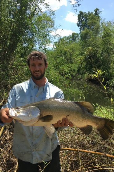 man holding a barramundi with water and thick vegetation behind