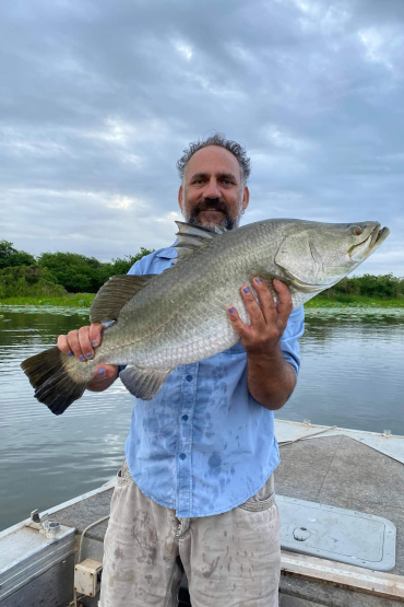 Colton Perna holding a large fish, standing in a boat on water, with bushes and other plants on the bank behind